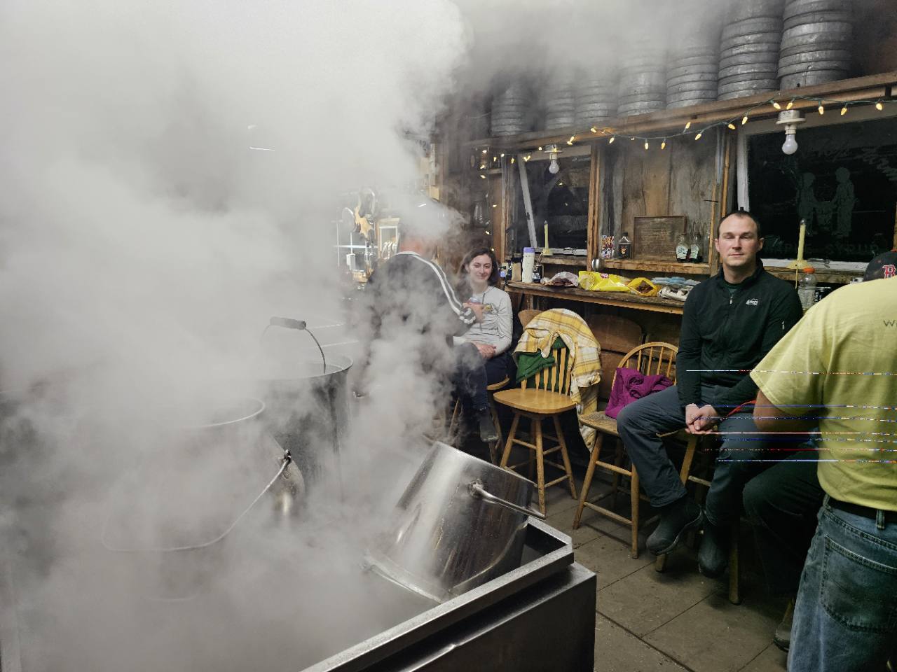 People sitting beside the steaming evaporator inside the Knolltop Farm sugarhouse